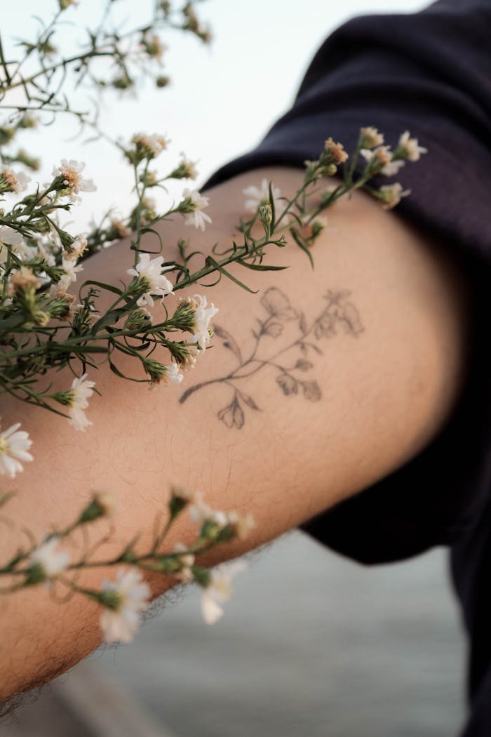 About Us A close-up of a floral tattoo surrounded by white flowers on a person's arm, evoking a natural and artistic vibe.