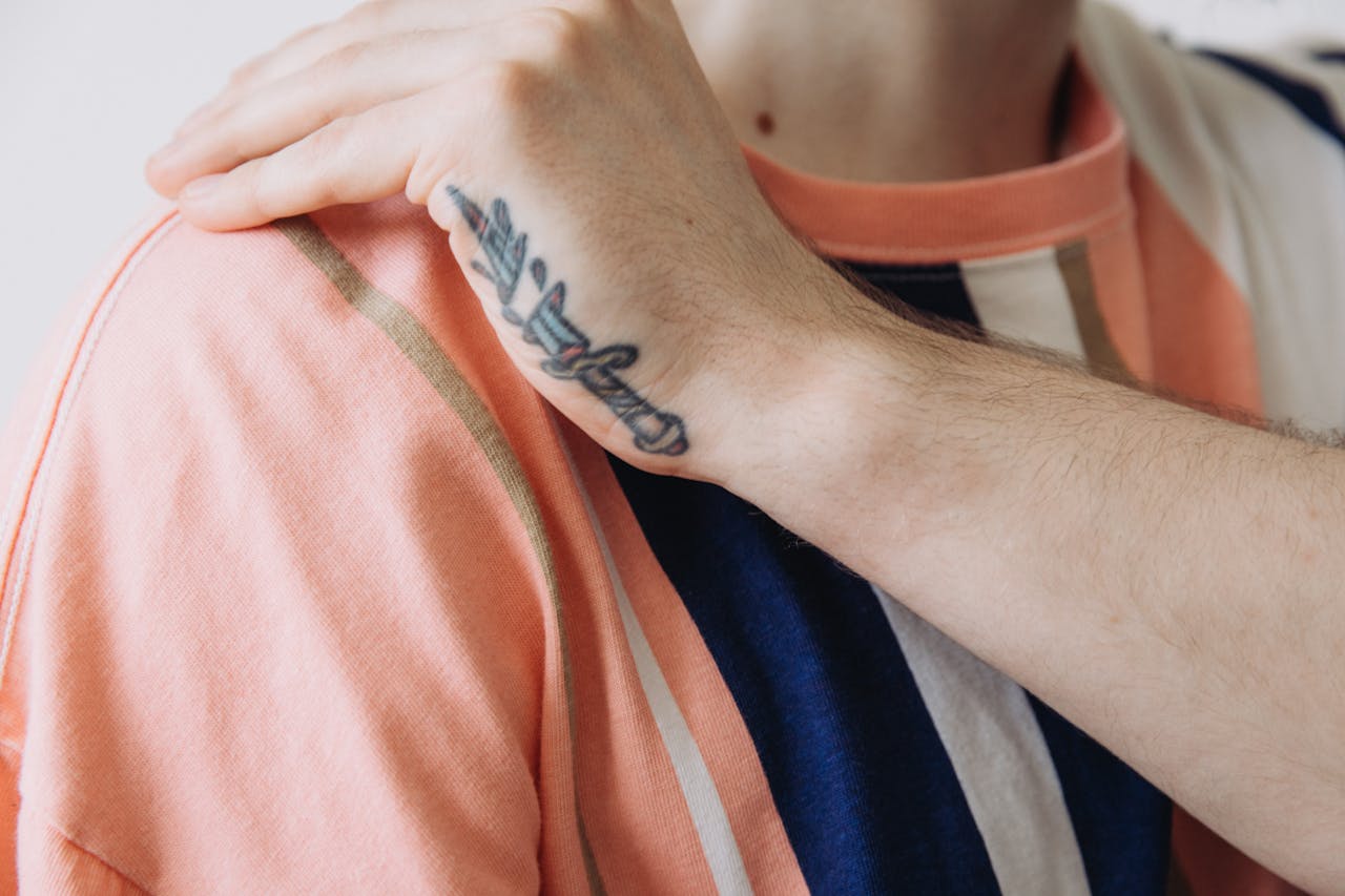 About Us A detailed close-up of a hand with a dagger tattoo resting on a striped shirt.